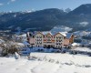 Hotel s bielou fasádou a balkónmi stojí na zasneženom svahu s horskou krajinou v pozadí.