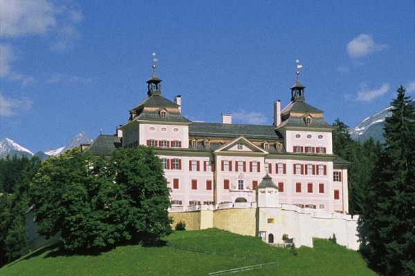 Historický zámok na kopci obklopený stromami s horami v pozadí.
