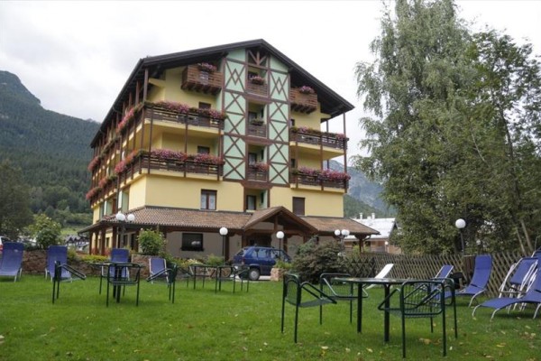 Štvorposchodový hotel s balkónmi a kvetmi obklopený záhradou so stoličkami.