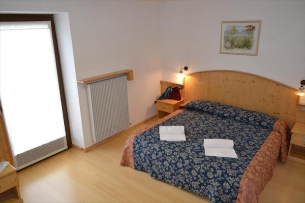 Spálňa s dvojlôžkom, nočnými stolíkmi, radiátorom a výhľadom na balkónové dvere.