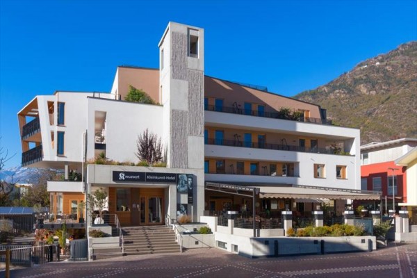 Moderný štvorposchodový hotel na úpätí hory s terasou a vonkajším sedením.  