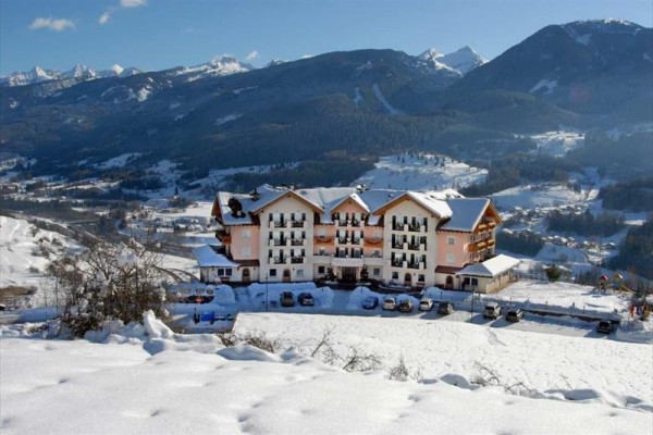 Hotel s bielou fasádou a balkónmi stojí na zasneženom svahu s horskou krajinou v pozadí.