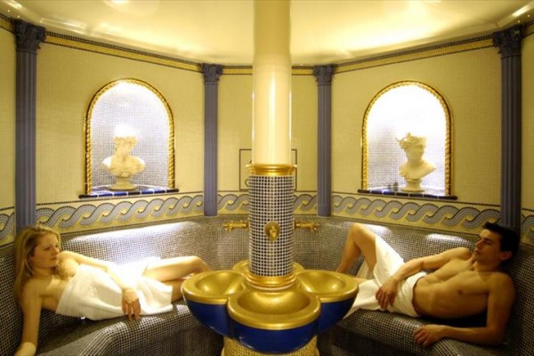 Dvaja ľudia relaxujú v luxusnej parnej kúpeľni s mozaikovými stenami a dekoratívnymi prvkami.