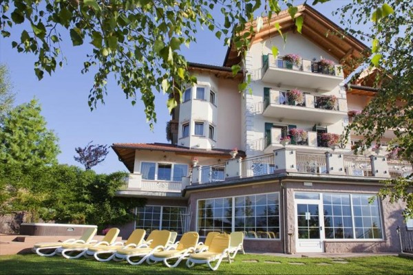 Budova s viacerými balkónmi a radom ležadiel pred ňou, obklopená zeleňou.