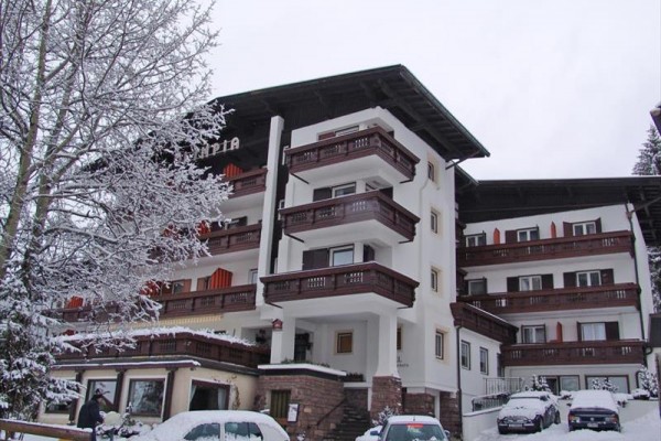 Štvorposchodový hotel so zasneženými balkónmi a zaparkovanými autami pred ním.