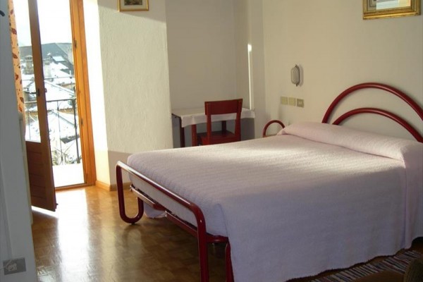 Svetlá izba s manželskou posteľou, červenou kovovou konštrukciou a otvorenými dverami na balkón.