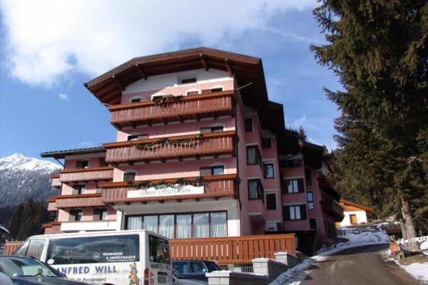 Horský hotel s drevenými balkónmi a bielym dodávkovým autom v popredí, zasnežené hory v pozadí.