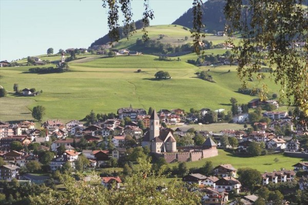 Stredoveký hrad obklopený domami v malebnej krajine s kopcami v pozadí.