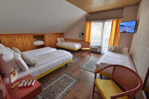 Izba s tromi lôžkami, TV, stoličkami a oknami s oranžovými závesmi.