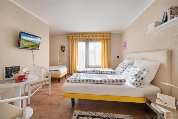 Svetlá izba s dvoma posteľami, oknom so žltými závesmi, televízorom a malým stolíkom so stoličkami.