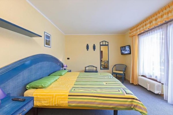 Izba s manželskou posteľou, žltým povlečením a televízorom na stene vedľa okna so závesmi.