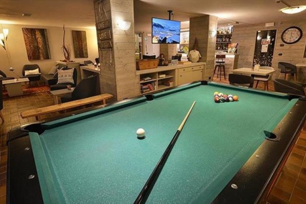 Interiér miestnosti s biliardovým stolom v popredí, televíziou a posedením v pozadí.