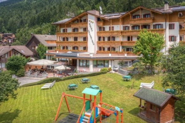 Hotel s terasou a detským ihriskom na trávniku obklopený lesom.