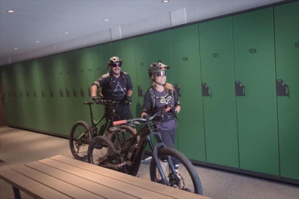 Dvaja cyklisti s prilbami tlačia bicykle popri rade zelených skriniek.