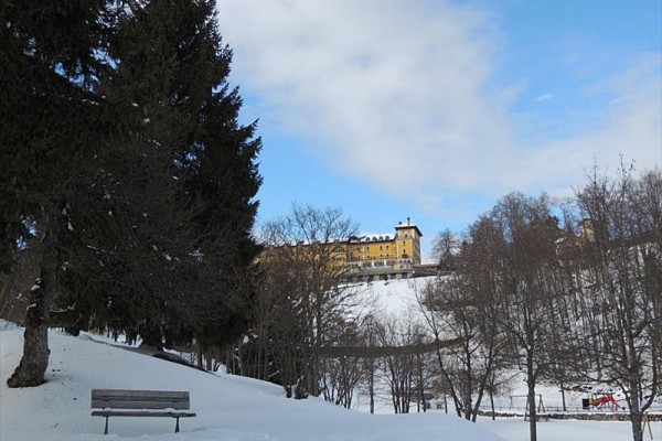 Snehom pokrytý park s lavičkou a v diaľke žltá budova na kopci.