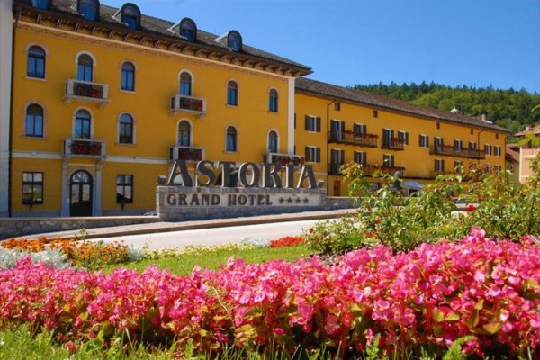 Žltá budova označená ako Grand Hotel Astoria s kvetmi v popredí.