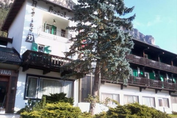 Hotel s balkónmi a zelenými okenicami obklopený horami a stromami.