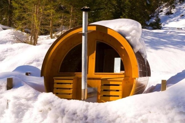 Drevená sudová sauna obklopená snehom v lesnom prostredí.