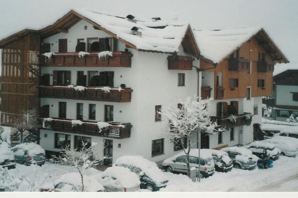 Zasnežený alpský hotel s balkónmi a zaparkovanými autami.