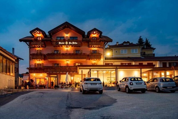 Večerný pohľad na osvetlený hotel s parkoviskom a autami pred ním.