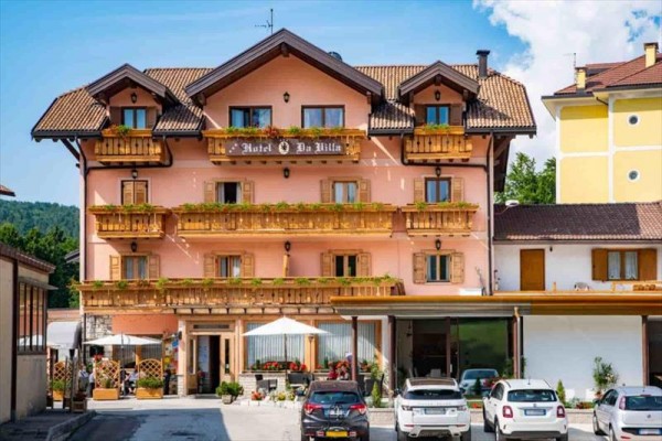 Trojposchodová ružová budova hotela s drevenými balkónmi a zaparkovanými autami pred ňou.  