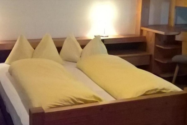 Manželská posteľ s dreveným rámom a žltou posteľnou bielizňou, vedľa nočného stolíka so svietiacou lampou.  