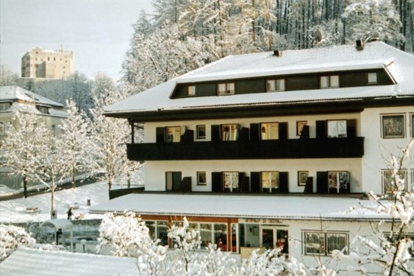 Budova s čiernymi balkónmi v zasneženej krajine, v pozadí les a zrúcanina hradu.
