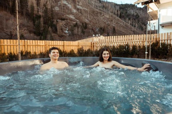 Dvaja ľudia relaxujú vo vonkajšej vírivke s dreveným plotom a horami v pozadí.