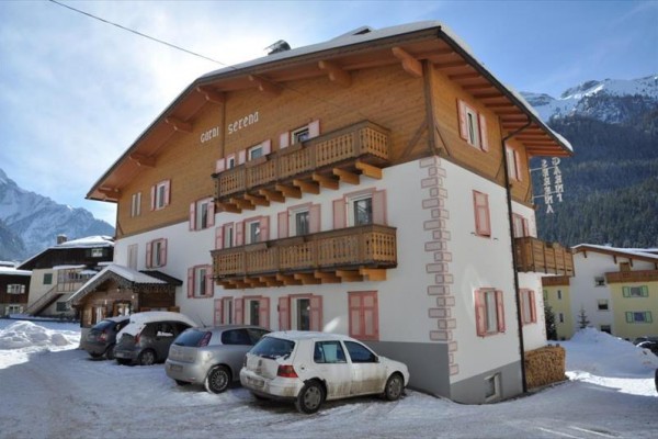 Horská ubytovňa s balkónmi, zasnežené parkovisko a autá, v pozadí hory.