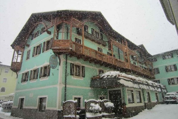Zasnežená budova s drevenými balkónmi a zelenou fasádou počas sneženia.  