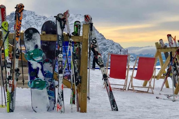 Lyže a snowboardy opreté o stojany na zasneženej zjazdovke s horami v pozadí.