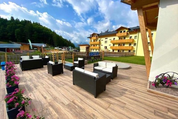 Terasa s ratanovým nábytkom, kvetmi a v pozadí žltý hotel pri lese.
