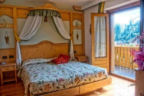 Spálňa s drevenou posteľou a otvorenými dverami na balkón s výhľadom na stromy.