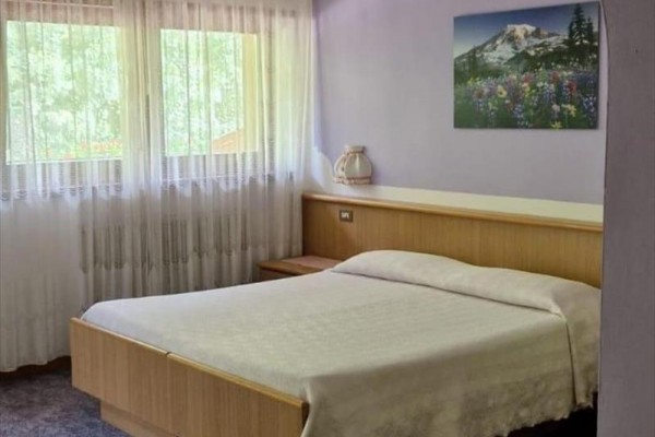 Jednoducho zariadená spálňa s manželskou posteľou, obrazom hory na stene a záclonami na oknách.