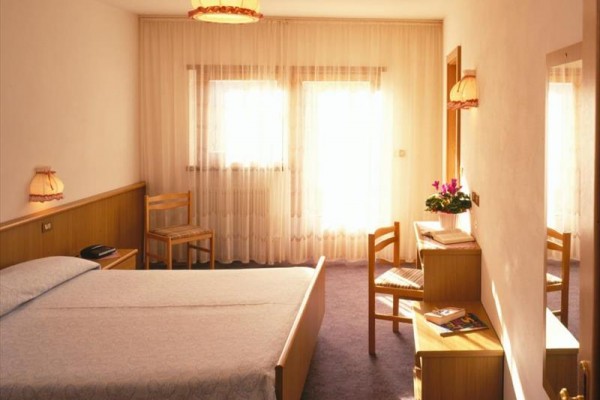 Svetlá hotelová izba s posteľou, nočnými stolíkmi, stoličkami a toaletným stolíkom pri okne so záclonami.