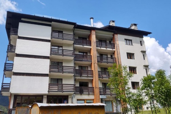Viacposchodová budova s balkónmi obklopená stromami pod modrou oblohou.