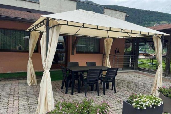 Altán s bielou plachtou a čiernym nábytkom na dláždenej terase.