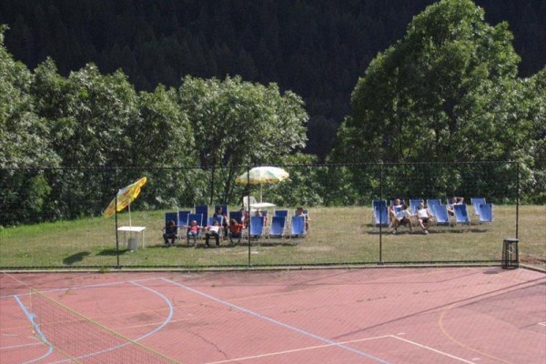 Ležadlá so slnečníkom na trávniku pri prázdnom športovom ihrisku obklopenom stromami.