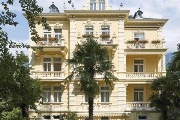 Žltá secesná budova s balkónmi, obklopená palmami a stromami.  