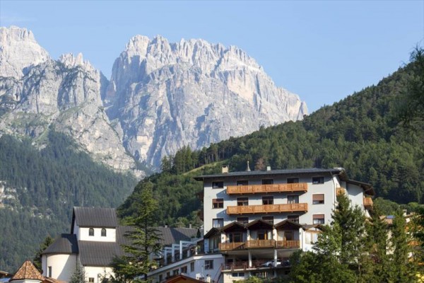 Hotel s drevenými balkónmi na úpätí zalesnenej hory so skalnatými vrcholmi.