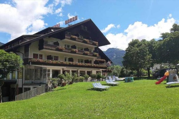 Hotel s balkónmi plnými kvetín, pred ktorým je rozsiahly trávnatý priestor s lehátkami a šmýkačkou.