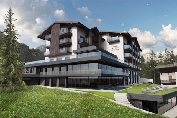 Moderný horský hotel so sklenenou fasádou obklopený lesmi a lúkou.