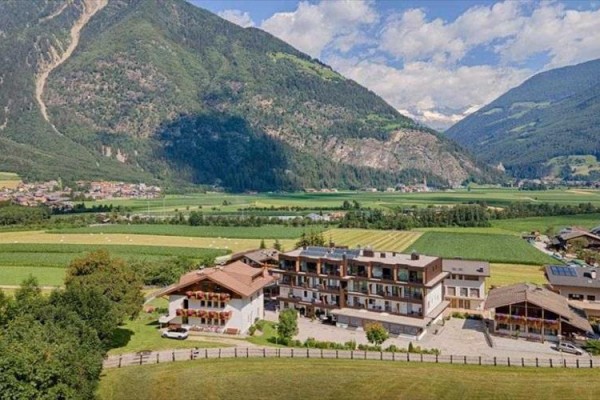 Hotel obklopený zelenými poli a horami na obzore pod modrou oblohou s oblaky.  