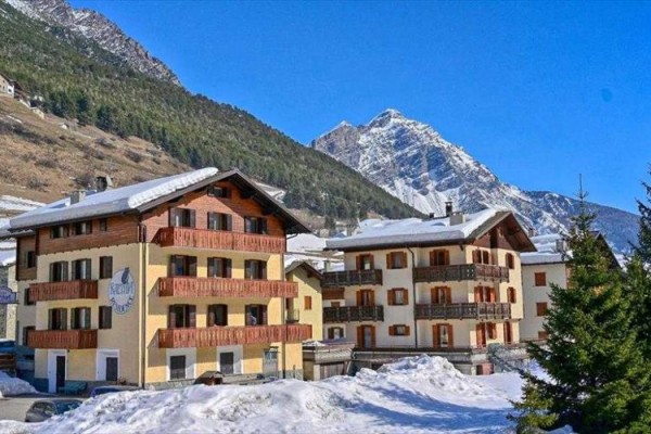 Horský hotel v zasneženej krajine s lesom a horami v pozadí.
