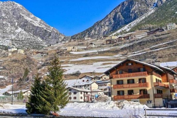 Alpská dedina s drevenými domami na svahu zasneženej hory pod modrou oblohou.