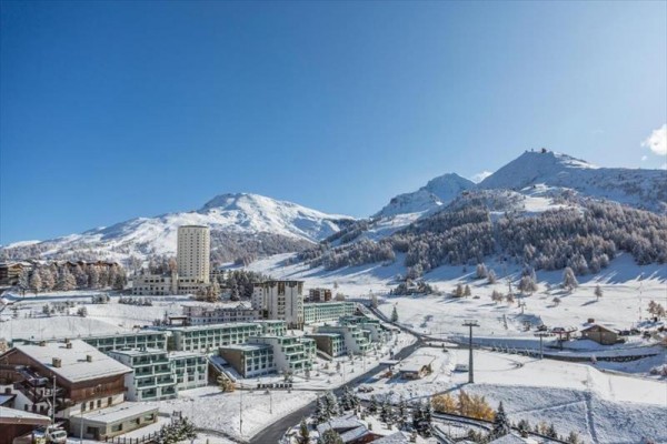 Zasnežená alpská krajina s lyžiarskym strediskom a horami v pozadí.