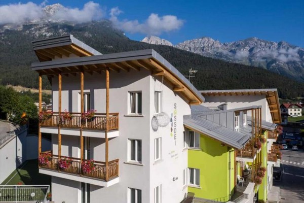 Moderný horský hotel so zelenou a bielou fasádou, v pozadí hory a obloha s oblačnosťou.