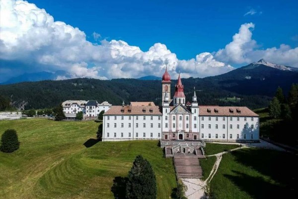 Historická budova s vežami na pozadí hôr a modrej oblohy s oblakmi.