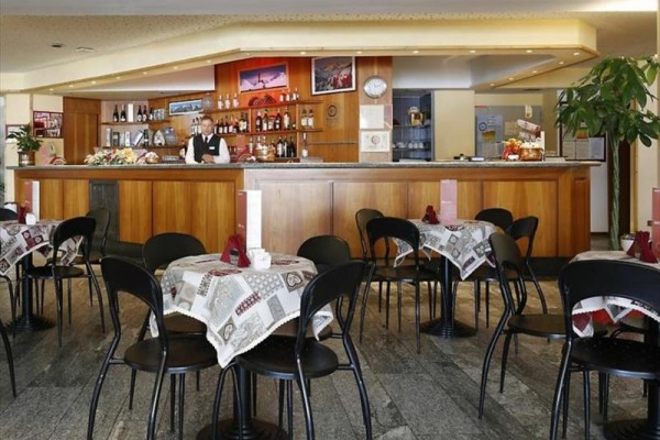Bar s dreveným pultom a stolmi s obrusmi, okolo ktorých je niekoľko stoličiek.