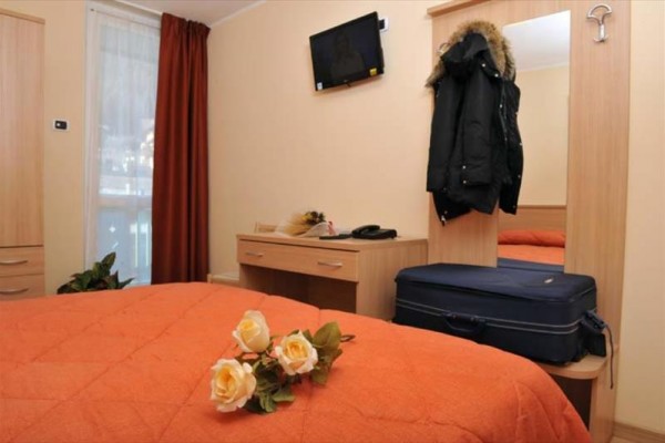 Hotelová izba s oranžovým prehozom na posteli, ružami a batožinou pri zrkadle.